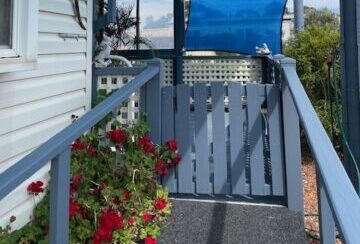 A narrow outdoor patio area with a grey railing and blue shade sails providing cover. Potted red flowers are in the foreground, and metal gecko wall art adorns the side of a white house. The sky is clear with some clouds in the background.