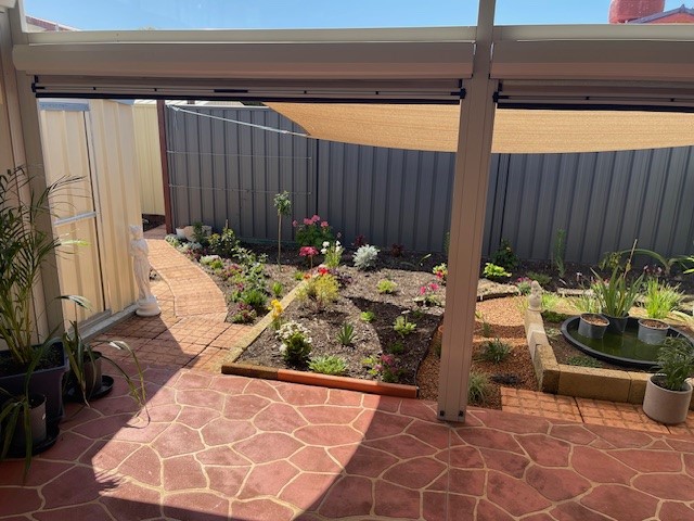 A tiled patio opens to a small garden with a variety of plants and flowers. A stone pathway curves through the garden, leading to a fenced backyard. There's a shaded area above the plants and a small fish pond with greenery on the right side of the garden.