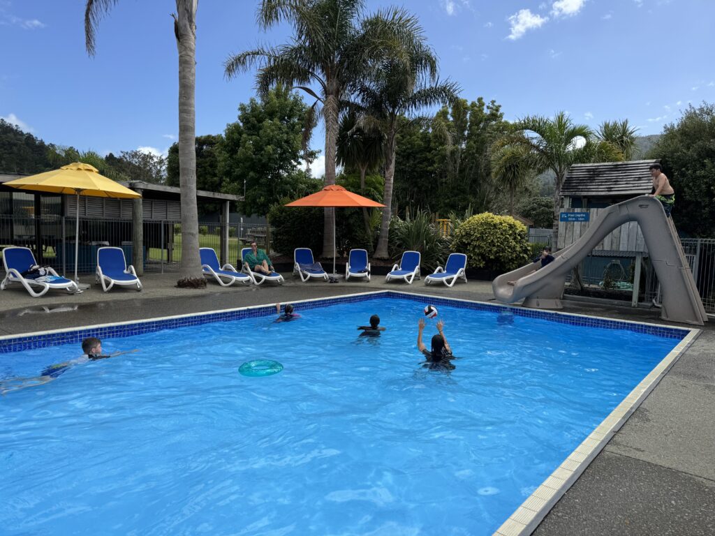 A swimming pool with several children playing in the water and using floating toys. Two individuals are at the poolside, and one child is going down a slide into the pool. There are lounge chairs with umbrellas around the pool, and lush greenery in the background.