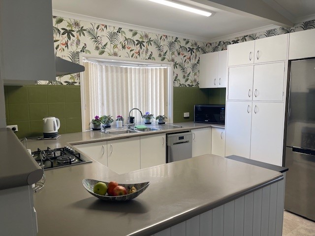 A modern kitchen featuring white cabinets, a stainless steel refrigerator, a dishwasher, and a microwave. A gas stove is located on the left with a kettle on the counter. The backsplash is green tile, and the upper walls have foliage-patterned wallpaper. A bowl of fruit sits on the island.