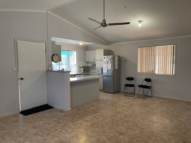 A spacious kitchen and dining area with a tiled floor. The kitchen has white cabinets, a refrigerator, a countertop with a microwave, and a wall clock. Two folded chairs are placed against the wall near a window with blinds. A ceiling fan hangs from the high ceiling.