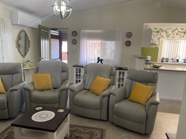 A cozy living room featuring four gray recliners with yellow cushions arranged around a dark wooden coffee table with a lace doily. The room is well-lit with natural light coming through lace-draped windows, and a kitchen area is visible in the background.