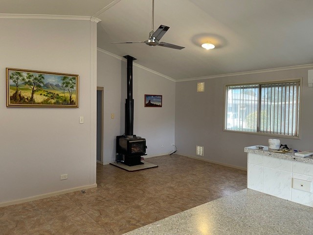 A spacious, sunlit room with light-colored walls and large windows. It features a ceiling fan, a wall-mounted painting, and a wood-burning stove in the corner. The floor is tiled, and there's a section of a countertop on the right side.