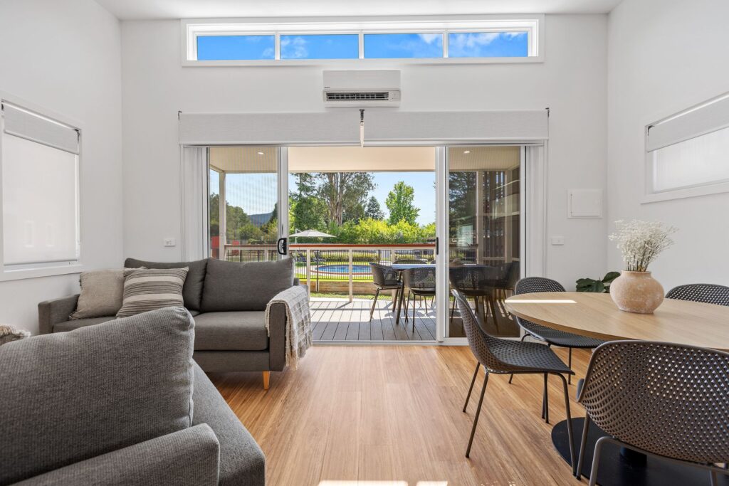 A modern living and dining area with wooden flooring and light-colored walls. The space features a gray sectional sofa, a dining table with black chairs, and large sliding glass doors leading to an outdoor deck with a view of trees and blue sky.