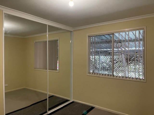 A small, unfurnished room with light beige walls. The room has a window on the right covered with vertical blinds and a mirrored sliding closet door on the left. A single ceiling light bulb illuminates the space. A rolled-up rug is on the floor.