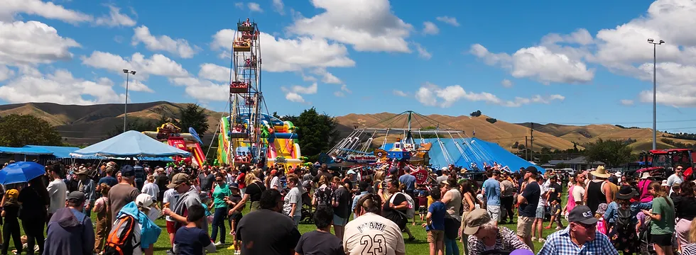 A lively carnival scene with a large crowd of people enjoying various attractions, including a Ferris wheel, inflatable slides, and a merry-go-round. The sky is blue with scattered clouds, and rolling hills are visible in the background.