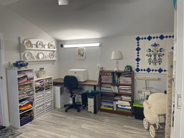 A well-organized craft room with shelves of fabric, a sewing machine on a wooden desk, wall-mounted shelves displaying decorative plates, a cushioned chair, and wooden bookshelves filled with crafting supplies. A white woven bassinet and a quilt adorn the space.