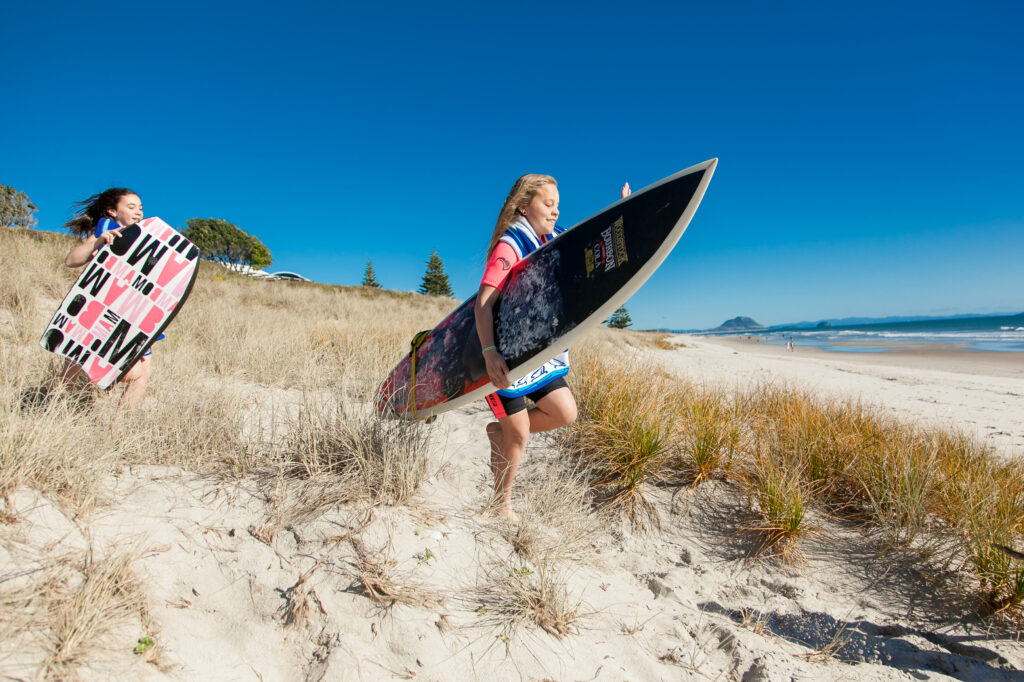 Two children carrying surfboards walk through sandy dunes towards a beach on a clear, sunny day. The child in the foreground holds a larger surfboard while the child behind carries a pink and black bodyboard. The ocean and distant mountains are visible. Showcasing premium beachfront access at Papamoa Beach