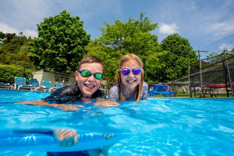 Two children smiling at the edge of an outdoor pool on a sunny day. They are wearing colorful swim goggles and holding onto a blue pool noodle. Having a laugh and enjoying water fun in Picton. Behind them, there are trees, poolside chairs, and a bright blue sky.