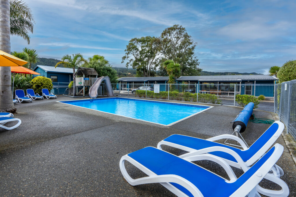 Outdoor swimming pool surrounded by blue and white lounge chairs and lush greenery. A gray water slide is visible on the left side, adjacent to the pool. A row of single-story buildings is in the background, with a scenic view of trees and hills under a partly cloudy sky.