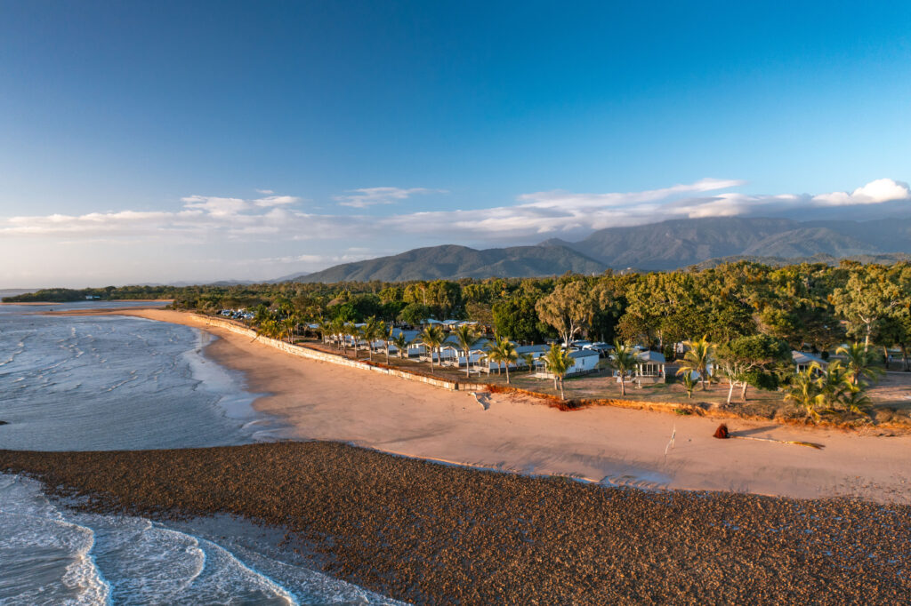 Aerial view of a picturesque beach with golden sand and calm waves meeting the shore. Lush green trees line the beach, with a few houses nestled among them. Behind the trees, a range of green hills extends into the blue sky, reminiscent of peaceful Rollingstone landscapes.