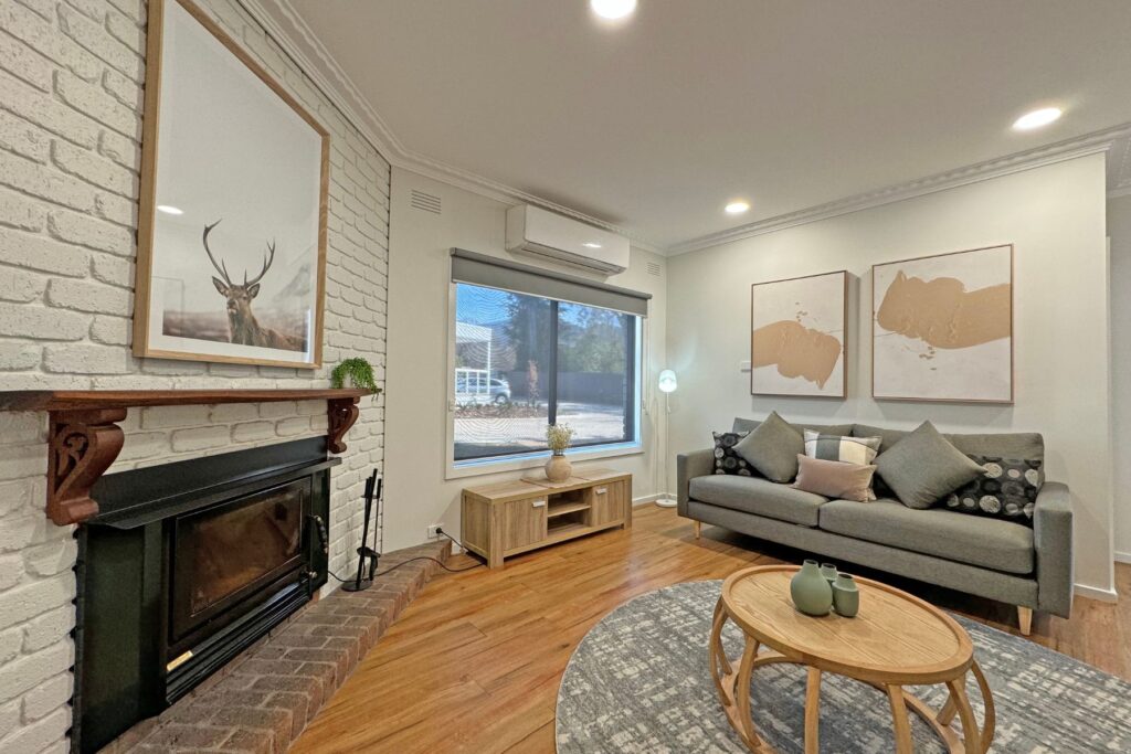 A modern living room at Retreat on Delany features a brick fireplace with framed deer artwork above it. A grey sofa with cushions sits against the wall adorned with abstract art. A round wooden coffee table and a TV stand are placed on a hardwood floor illuminated by recessed lighting.