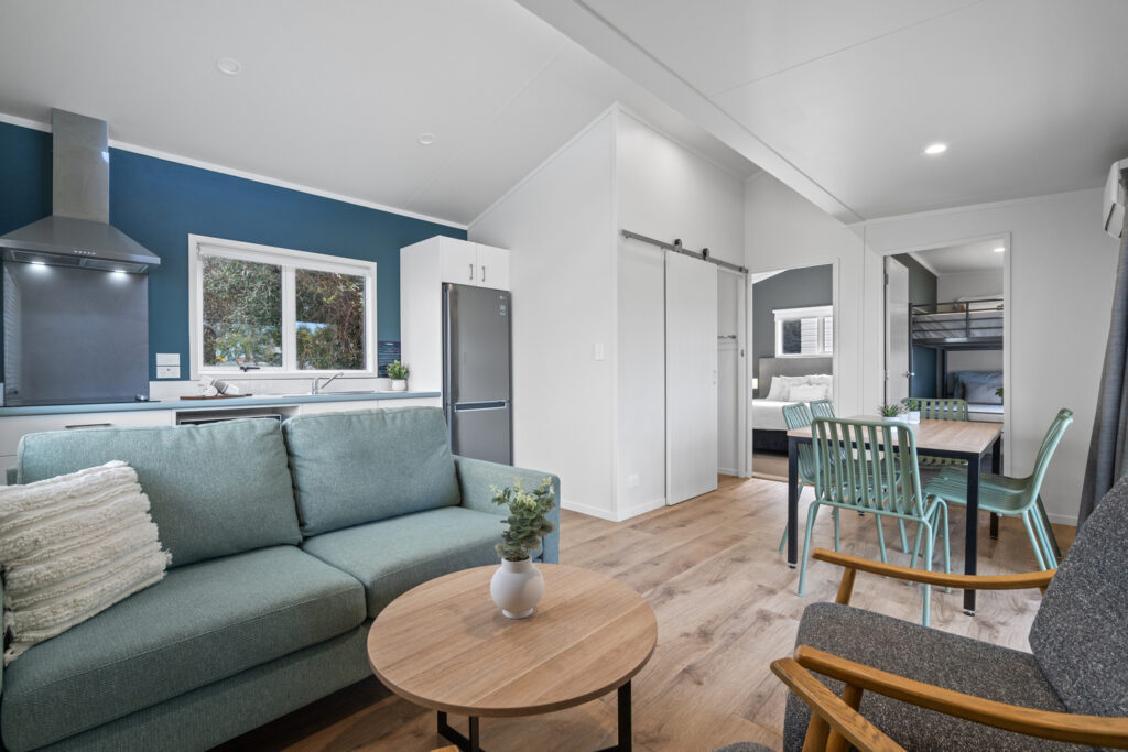 A new, modern, open-concept living space features a kitchen with dark cabinets and a stainless steel refrigerator. The living area boasts a teal sofa and wooden coffee table. A dining table with green chairs sits near a sliding door leading to a bedroom and bunk room.