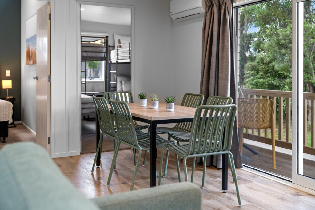 A dining area with a new wooden table and six green metal chairs. Three small potted plants are on the table. A sliding glass door leads to a deck outside, and an open doorway reveals a room with bunk beds. A green sofa is partially visible in the foreground.