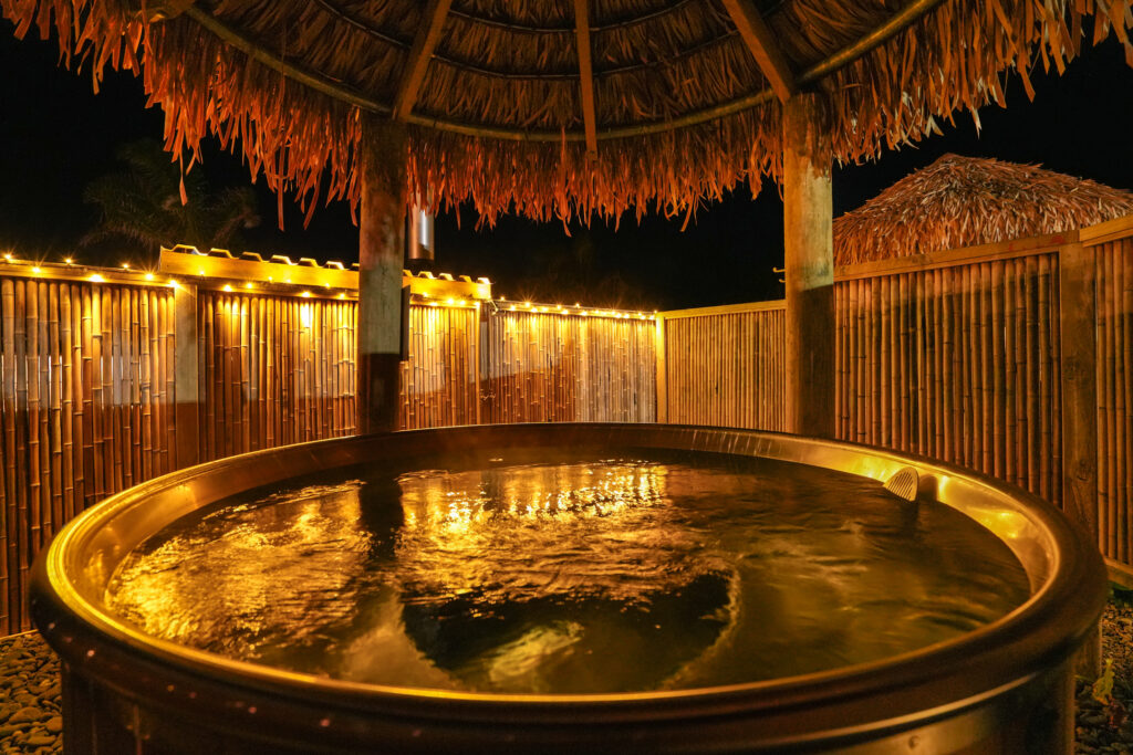 A luxurious outdoor hot tub at night, encircled by a bamboo fence and softly lit with string lights. The thatched roof creates a cozy, tropical ambiance. The water reflects the warm glow of the lights, enhancing the serene and relaxing atmosphere.