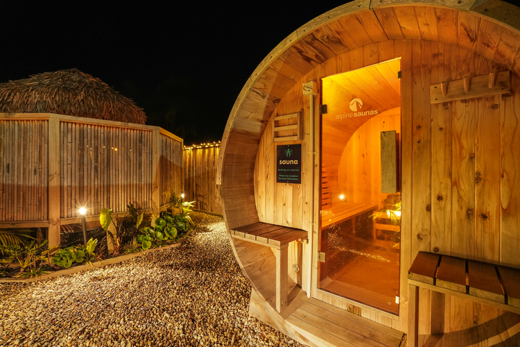 A wooden barrel-shaped outdoor sauna with warm interior lighting is set in a neatly landscaped area with small pebbles and plants. A circular window and a glass door with a visible sign enhance the rustic ambiance. A thatched-roof structure is nearby.