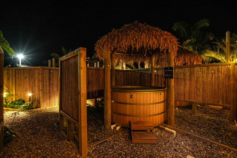 A wooden hot tub under a thatched roof is shown in an outdoor setting at night. It is surrounded by a bamboo privacy fence and illuminated by warm lights, creating a cozy and inviting atmosphere. Trees and additional lights are visible in the background.