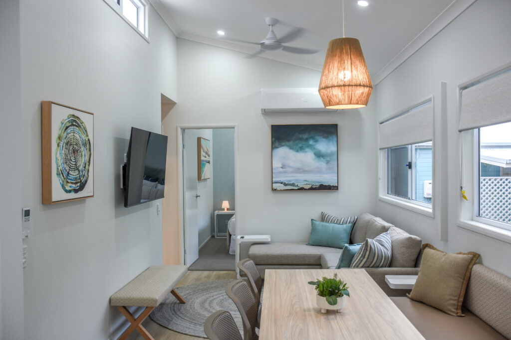 A modern, well-lit living area with a dining table in the foreground. The room includes a beige sectional sofa, wall-mounted TV, and serene North Star artwork. Several windows allow natural light in, and a woven pendant light hangs from the ceiling. A bedroom entrance is visible.