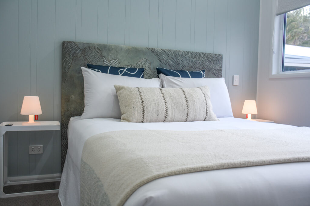 A neatly made bed with white linens and a large decorative pillow, featuring a gray textured headboard and blue accent pillows, sits between two bedside tables each holding a lit lamp. The walls are painted light gray, resembling the cool hue of the North Star, and a window in the corner brings in natural light.