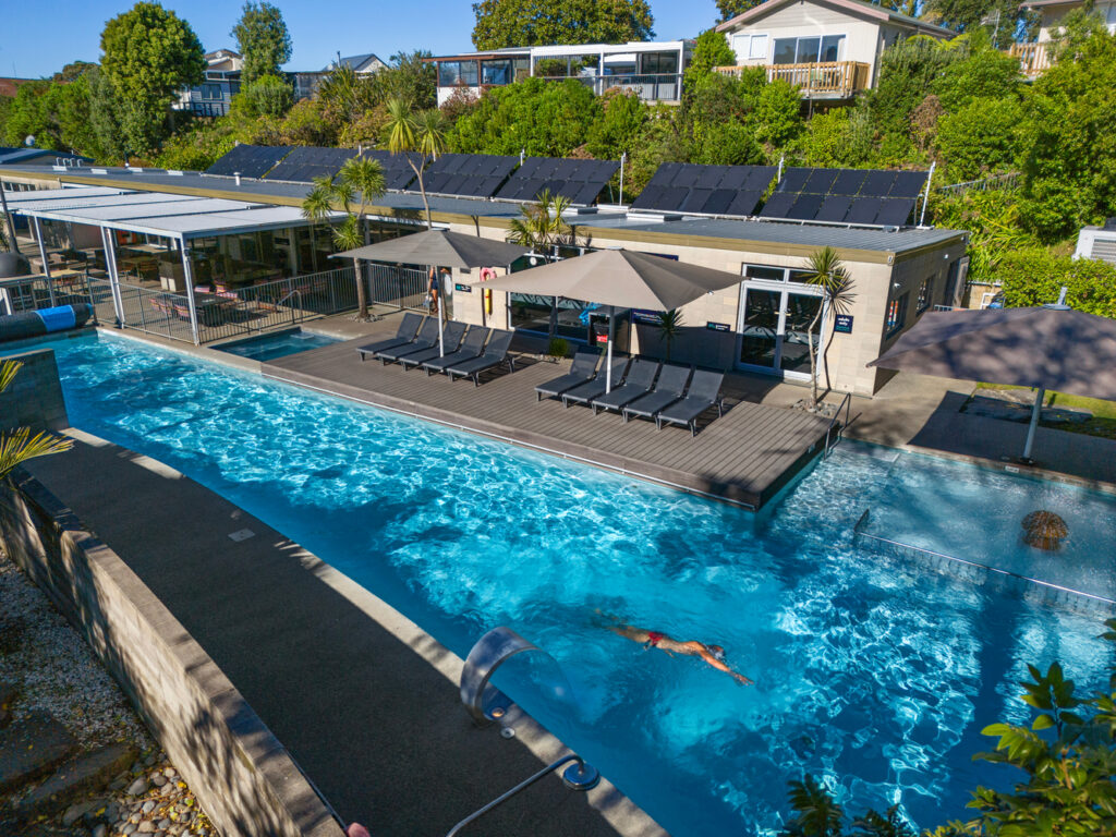 A person swims in an outdoor rectangular pool bordered by a wooden deck with several lounge chairs and large umbrellas. The modern building in the background features solar panels on the roof. Lush greenery and residential houses are visible beyond the pool area.