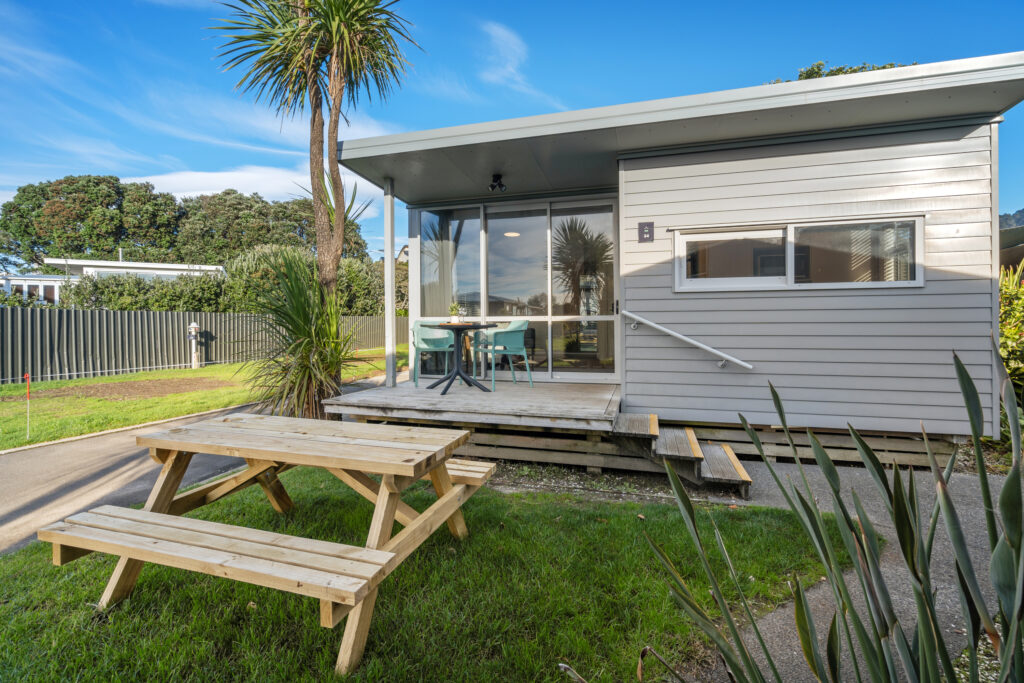 A small, modern grey cabin with a covered porch and new glass sliding doors features an outdoor dining area. A wooden picnic table is situated on the grass in the foreground. Palm trees and a clear blue sky create a tranquil backdrop.