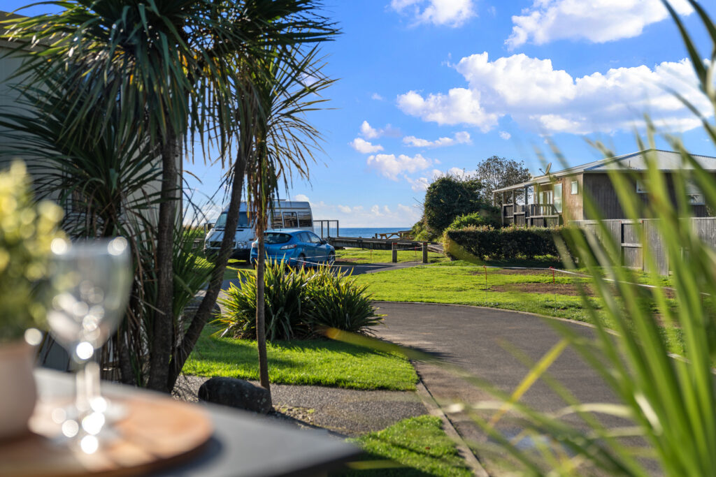 A scenic view of a new residential area with a clear blue sky. Palm trees frame the image, and a few parked vehicles, including a camper van, are visible. A pathway leads towards the ocean in the distance. A blurred foreground shows a table set with a wine glass.