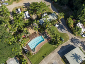 Aerial view of Cairns Cool Waters resort's pool area surrounded by several buildings and lush vegetation. The kidney-shaped pool is adjacent to a smaller rectangular pool. Numerous palm trees and green spaces are visible, along with parked vehicles and recreational vehicles.