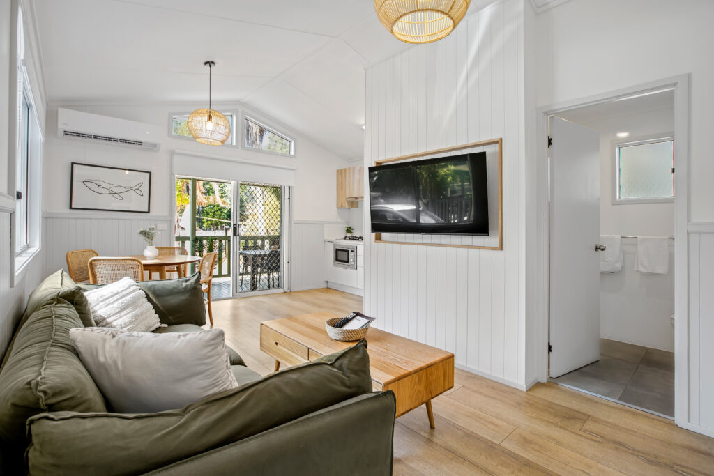 A modern, cozy living area with a green sofa and white pillows, a wooden coffee table, a large TV mounted on the white paneled wall, and a dining table with chairs near a sliding glass door. The kitchen and a partially visible bathroom are in the background. Natural light fills the space.