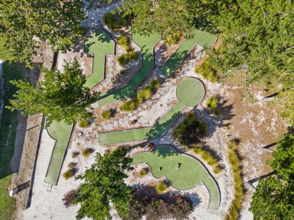 Aerial view of a mini-golf course during spring in Ohiwa, with winding green pathways and putting areas surrounded by blossoming trees and shrubs. Pathways are bordered by stones, and there are sandy sections interspersed among the course.