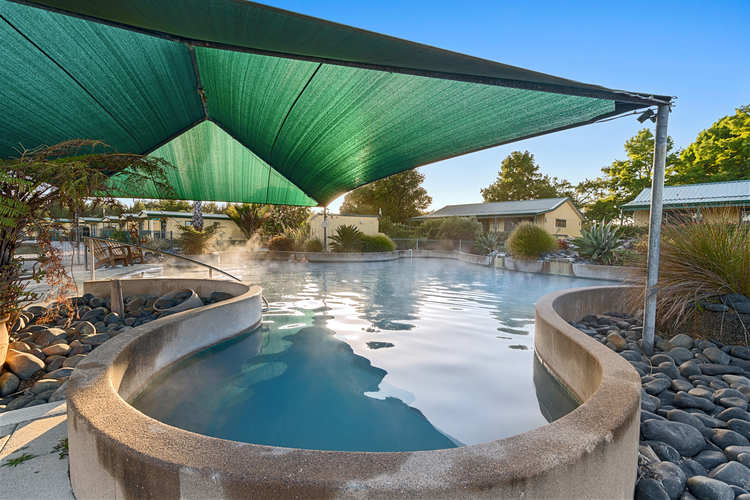 A serene outdoor hot spring pool with steam rising from the water. The pool is shaded by a large green tarp. Surrounding the pool are rocks and lush greenery. In the background, there are small buildings and trees against a bright blue sky.