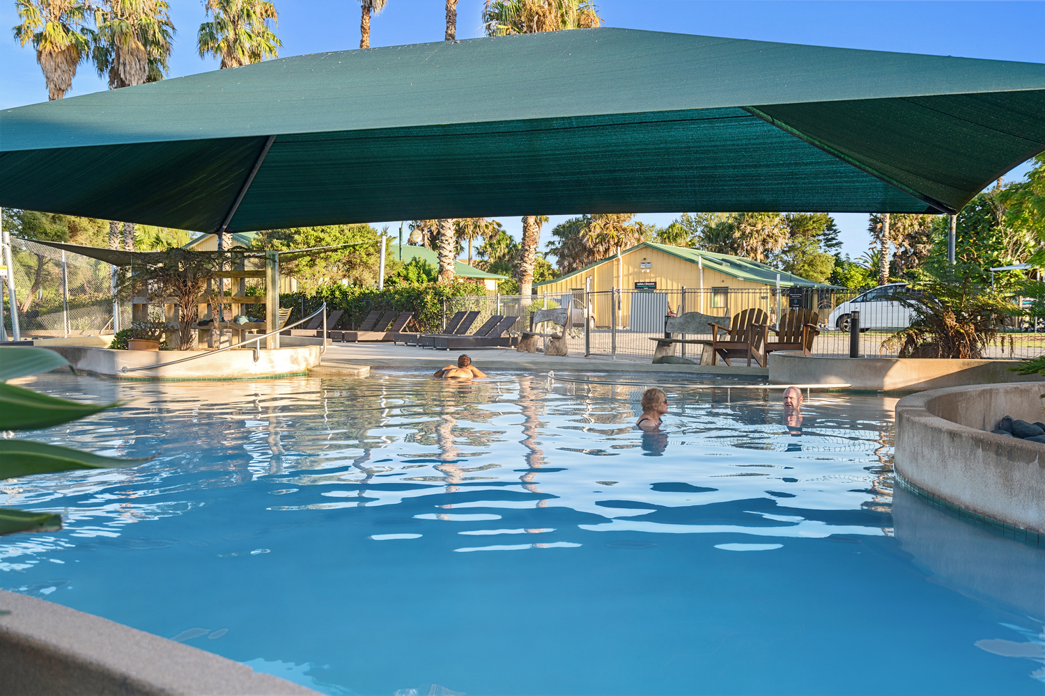 A serene outdoor mineral hot pool, reminiscent of spring in Ohiwa, is shaded by a large green canopy. Some people are swimming and relaxing in the water. In the background, there are lounge chairs, a small building, and palm trees against a blue sky.