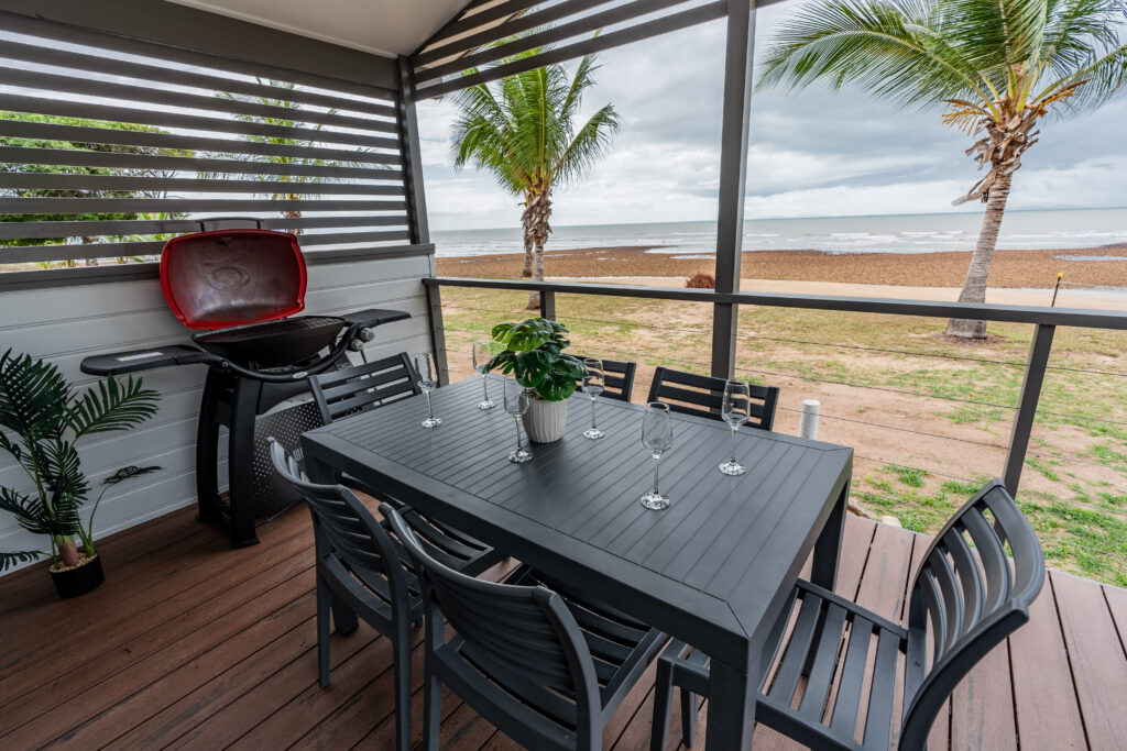 A covered outdoor deck overlooking a beach, featuring a black dining table with five chairs, a potted plant, and several empty wine glasses. A red Rollingstone barbecue grill is placed against the railing, and palm trees are visible in the background.