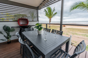 A patio with a dark gray dining table set for six, featuring wine glasses and a potted plant, exudes a Rollingstone vibe. A barbecue grill stands to the side, offering the perfect blend of relaxation and adventure as it overlooks a beach with palm trees under a cloudy sky.