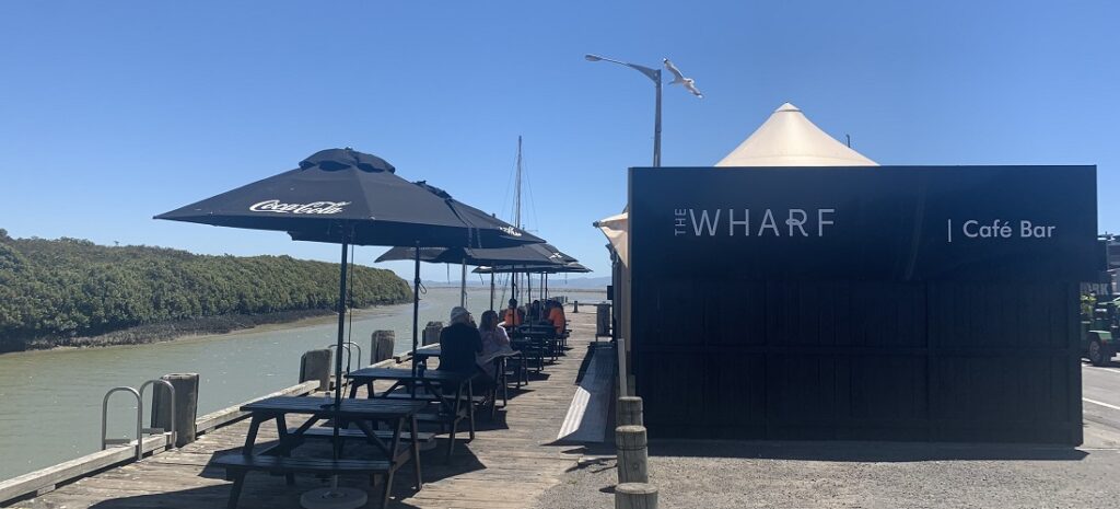 An outdoor seating area next to a river with several picnic tables and umbrellas. People are sitting at the tables, and a black sign to the right reads "THE WHARF | Café Bar." Mangroves and a clear blue sky enhance the serene ambiance, capturing the essence of Spring in Ohiwa.