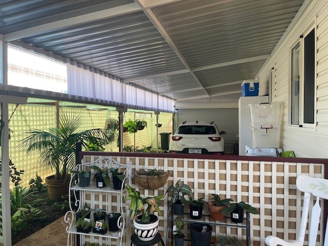 A covered patio area with a white SUV parked on a concrete driveway. The patio features potted plants on a multi-tiered stand, hanging planters, and lattice fencing. The area is sheltered by a corrugated metal roof.