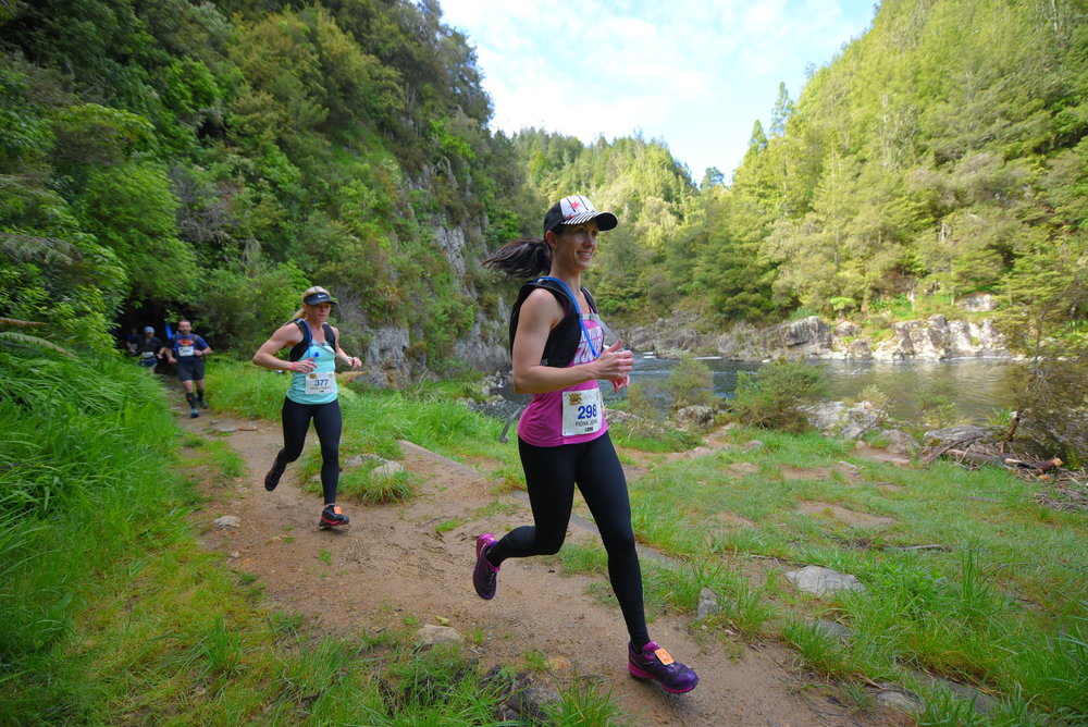 Two women run along a scenic riverside trail surrounded by lush greenery. Both are wearing athletic gear and race numbers, with the woman in the foreground holding a water bottle and smiling. The sun shines on the verdant landscape, creating a vibrant, cheerful atmosphere.