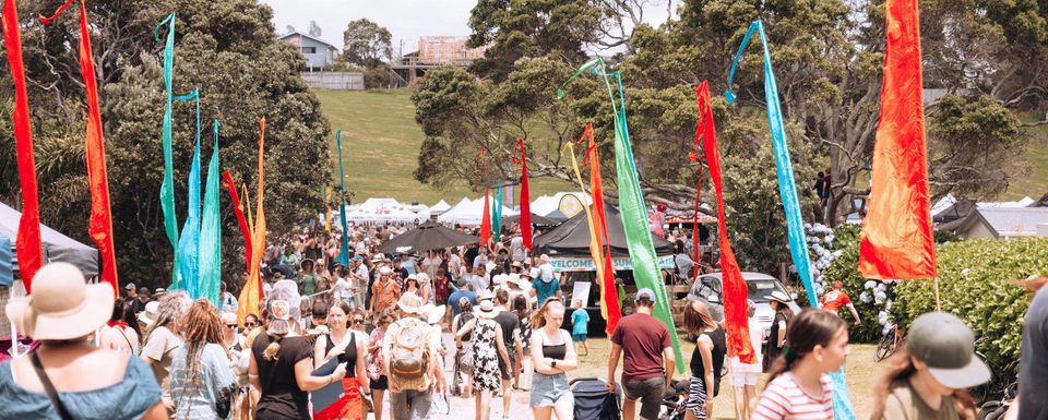 A vibrant outdoor festival with a large crowd of people walking among colorful flags and banners. Trees, tents, and booths are visible in the background, with some people wearing hats and backpacks. The atmosphere is lively and bustling.