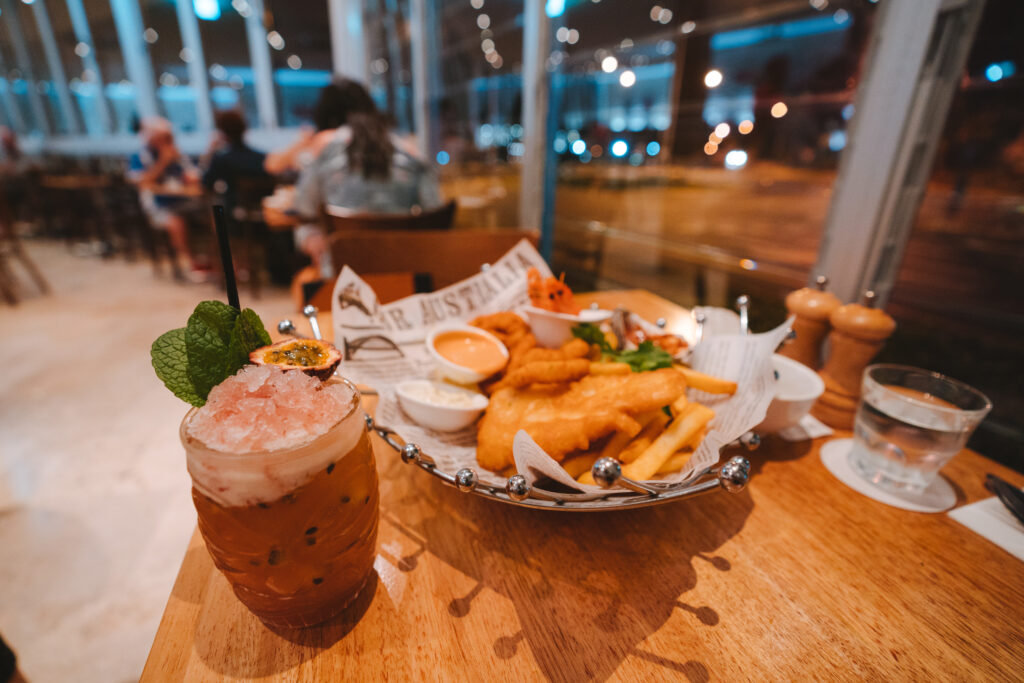 Discover the essence of local cuisine at a wooden table adorned with a mint and passion fruit cocktail beside a platter of fish and chips, complete with dipping sauce and a small salad, all set in a bustling restaurant ambiance.