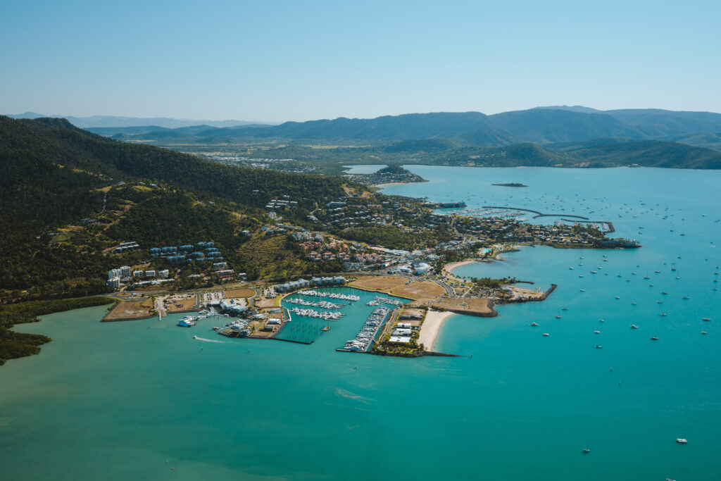 A scenic aerial view of a coastal town with a marina, surrounded by turquoise waters, invites exploration. Boats scatter the water while lush green hills and distant mountains frame the landscape under a clear blue sky—a perfect backdrop for discovering local cuisine.