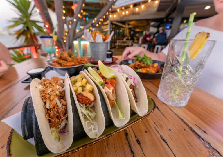 A table at a restaurant showcases local cuisine with a close-up of various tacos in a holder, accompanied by a drink garnished with pineapple. People savor the flavors amid string lights and a cozy, vibrant atmosphere.