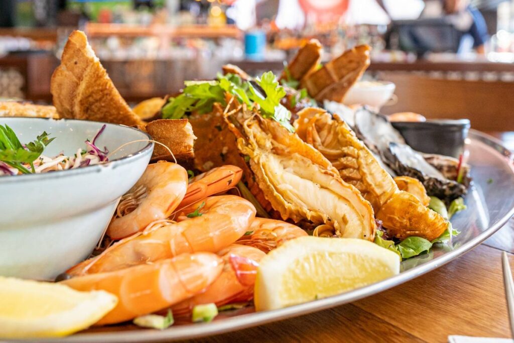 A platter of assorted seafood featuring shrimp, lobster tails, oysters, and lemon wedges, garnished with fresh herbs invites you to discover local cuisine. A bowl of salad is visible in the foreground, all set on a wooden table in a lively restaurant setting.