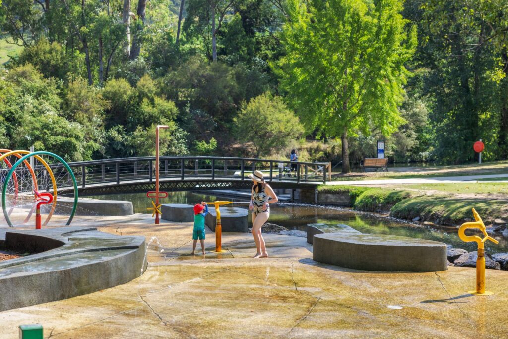 A child and adult play at  Bright splash park with water features and sprays is one of the best things to do in Bright. A bridge is in the background, surrounded by trees and greenery under a clear blue sky.