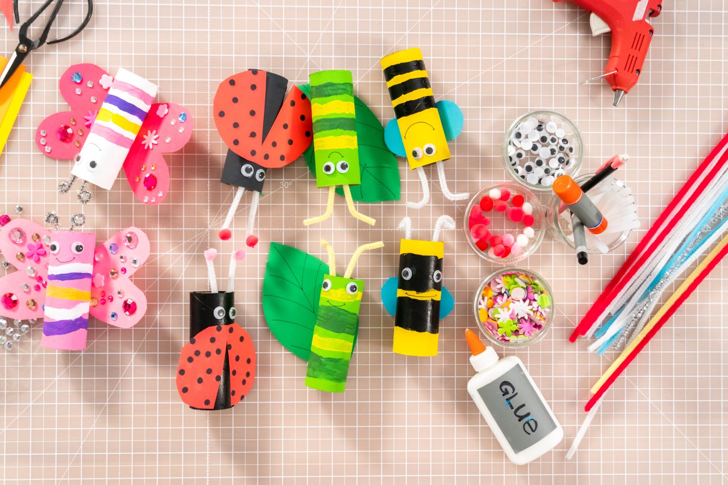 A collection of colorful, handmade insect crafts made from recycled toilet paper rolls is displayed. There are butterflies, ladybugs, and bees alongside craft supplies like googly eyes, glue, and pipe cleaners on a grid-patterned surface.