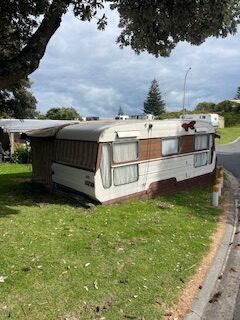 A vintage caravan with a striped awning is parked on a grassy area near a road. Several other caravans are visible in the background under a cloudy sky.