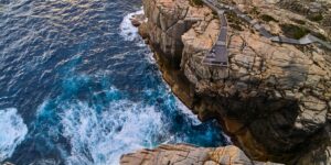 Aerial view of a rocky coastline with a wooden boardwalk extending along the cliff edge. Waves crash against the rocks, creating white foam on the blue ocean water below. Nearby, caravans dot the landscape, offering travelers a dramatic view of the rugged seascape from their temporary homes.