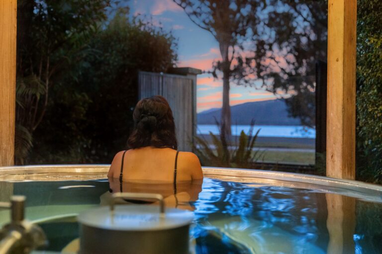 A person with dark hair is relaxing in an outdoor hot tub, facing a scenic view of a lake or sea. The sky is painted with the colors of a sunset, and there are trees surrounding the area.