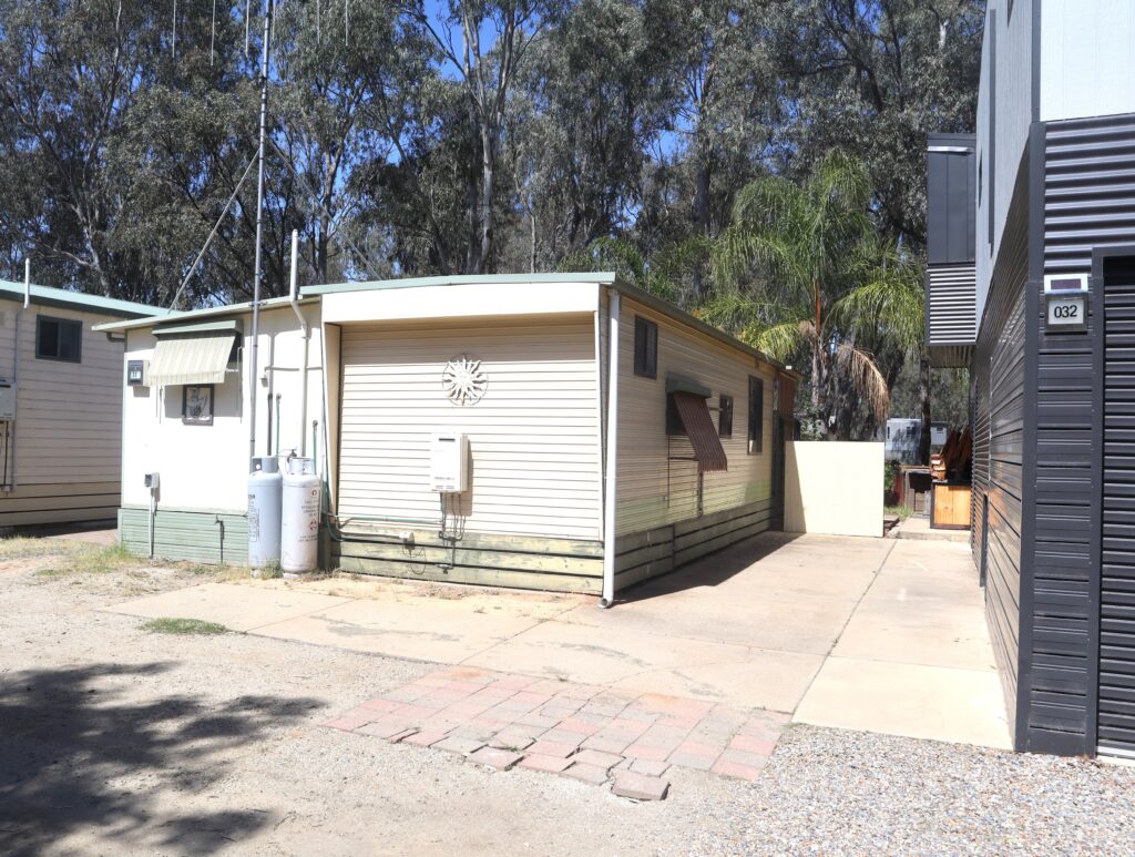 A small, light-colored mobile home with a flat roof is situated on a gravel driveway. There are trees and another similar structure nearby, with utility meters visible on the side. A patch of sunlight brightens the entrance area.