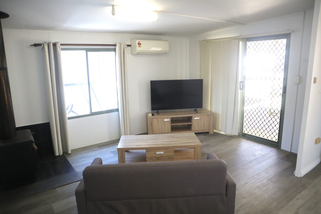 A cozy living room featuring a gray couch facing a flat-screen TV on a wooden stand. The room has a fireplace, light-colored curtains, a large window, and sliding glass doors leading outside. The flooring is wood laminate.