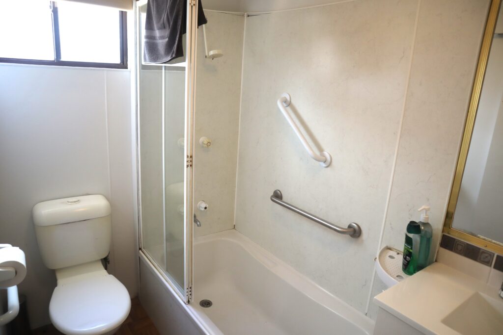 A bathroom with a white toilet, a bathtub with grab bars, and a glass shower door. The sink has a soap dispenser, and a towel hangs over the shower door. A window provides natural light.