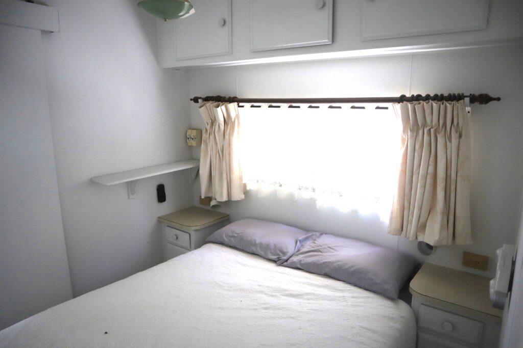 A cozy bedroom with a double bed covered in white linens and two gray pillows. The room has white walls and cabinetry, a small window with cream curtains above the bed, and side tables on each side. Soft lighting fills the space.